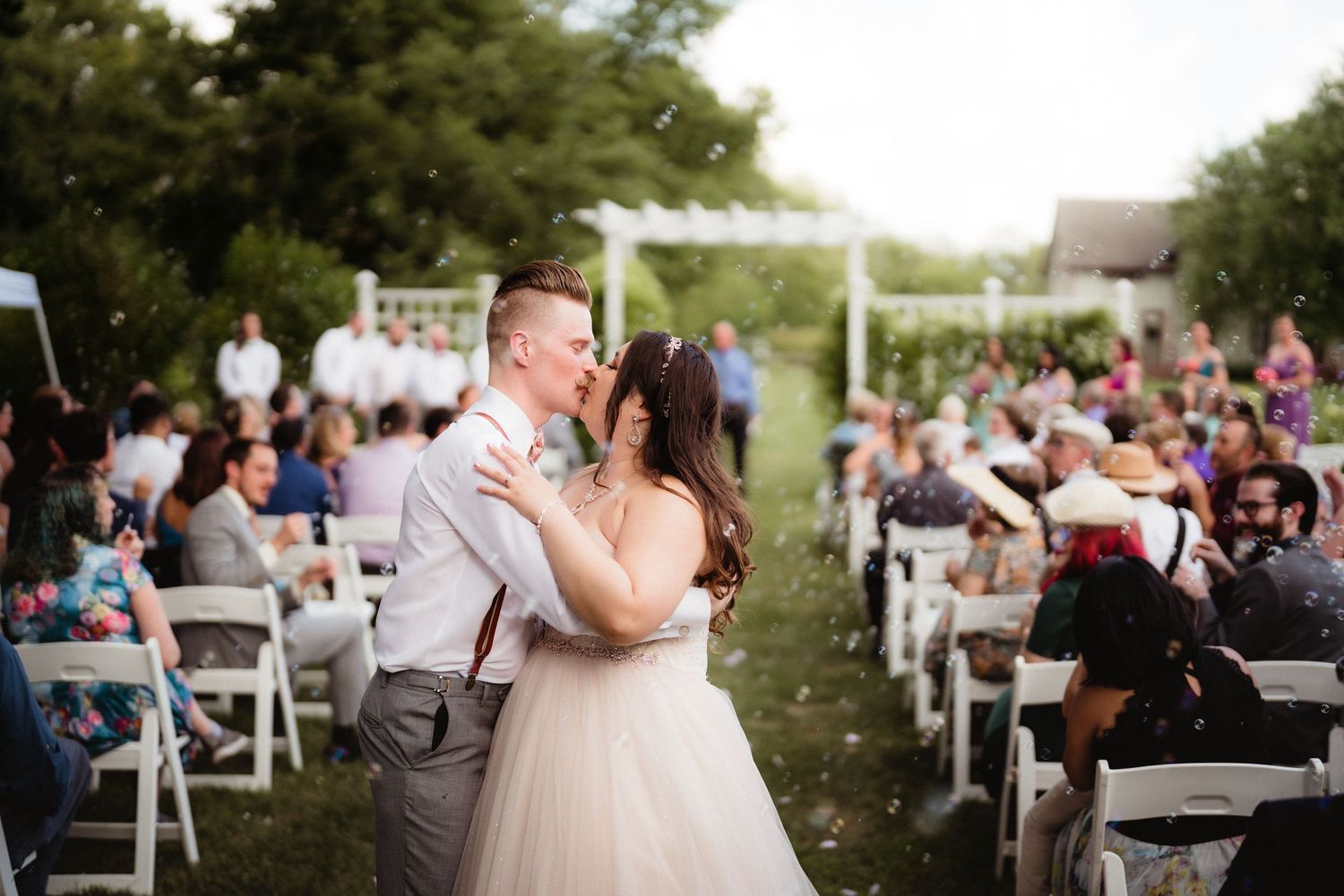 Couple embraces during outdoor wedding ceremony with white chairs and guests in background.