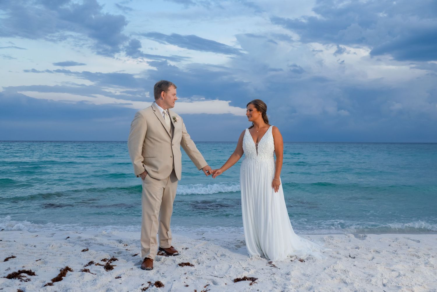 Newlyweds holding hands while standing on white sand beach at dusk with turquoise ocean backdrop.