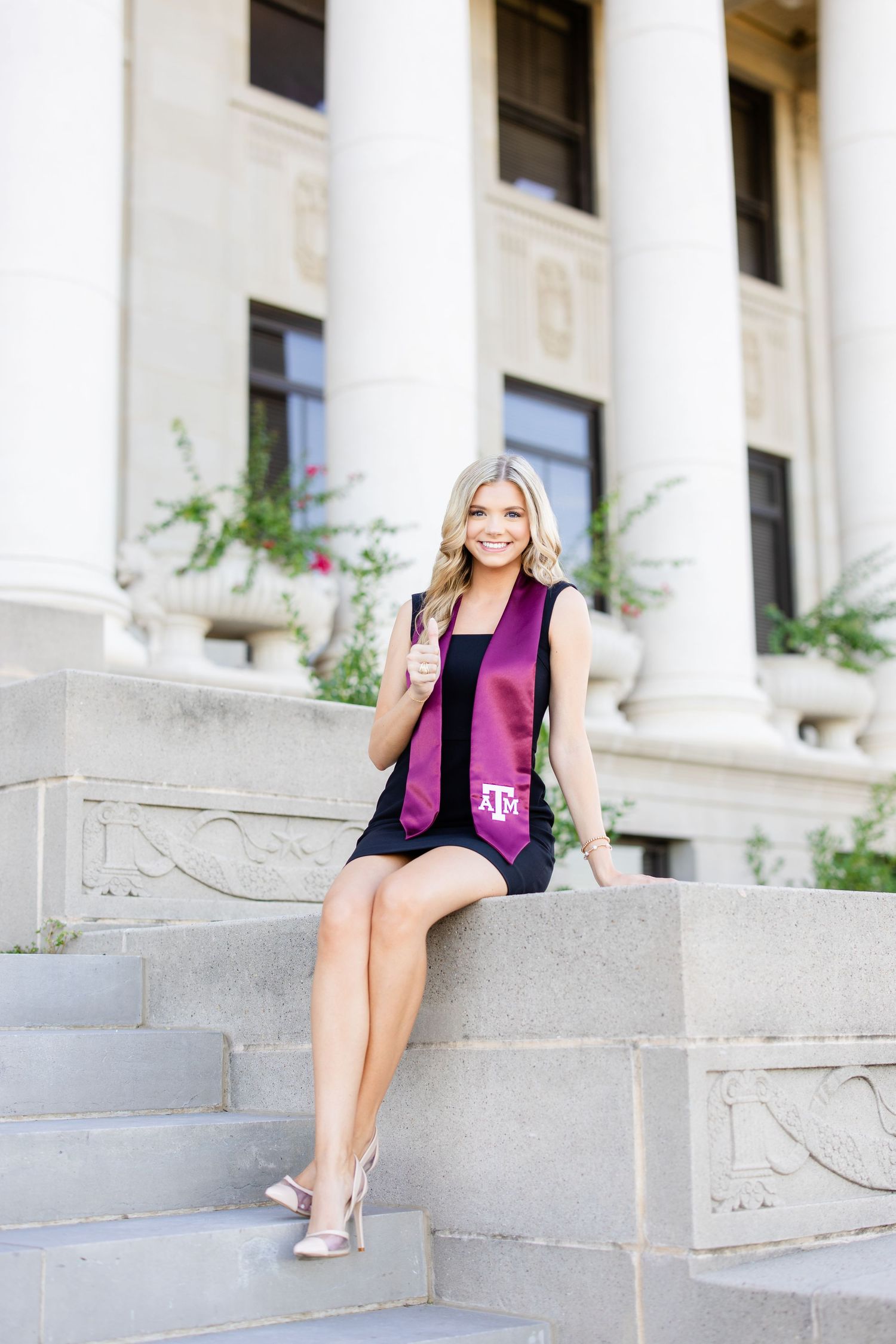 Graduate in black dress and maroon stole sitting on stone steps of campus building.
