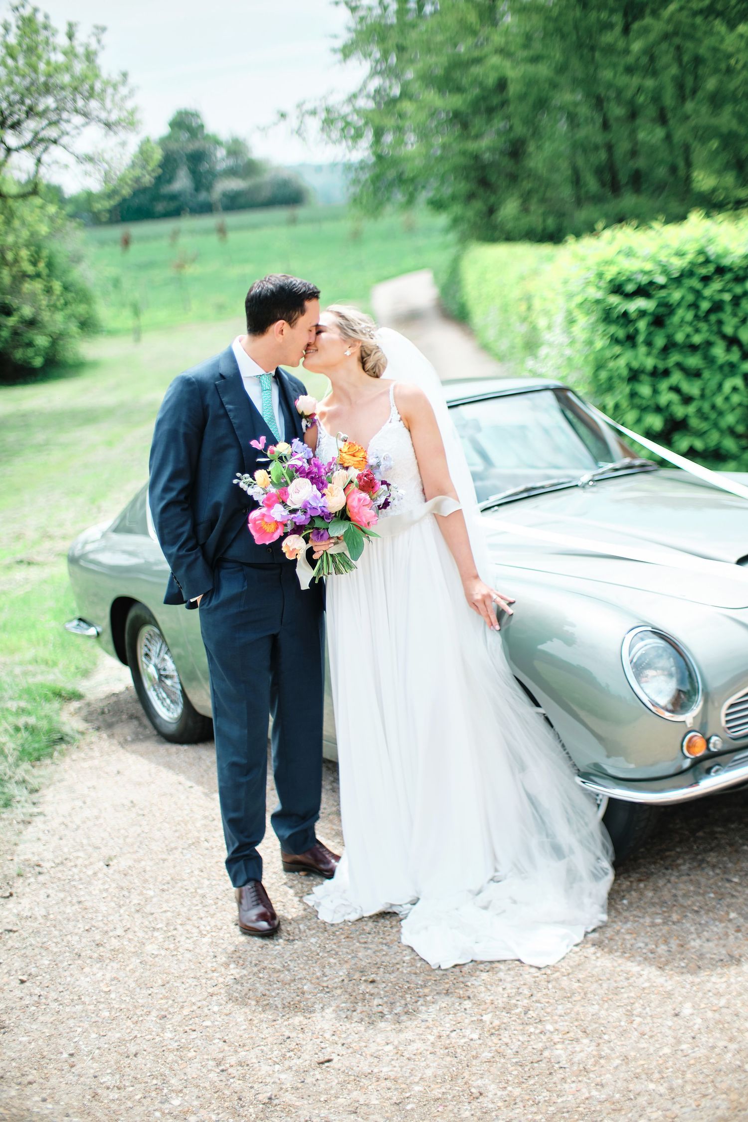 A couple poses with their vintage silver car amidst lush greenery on their wedding day.