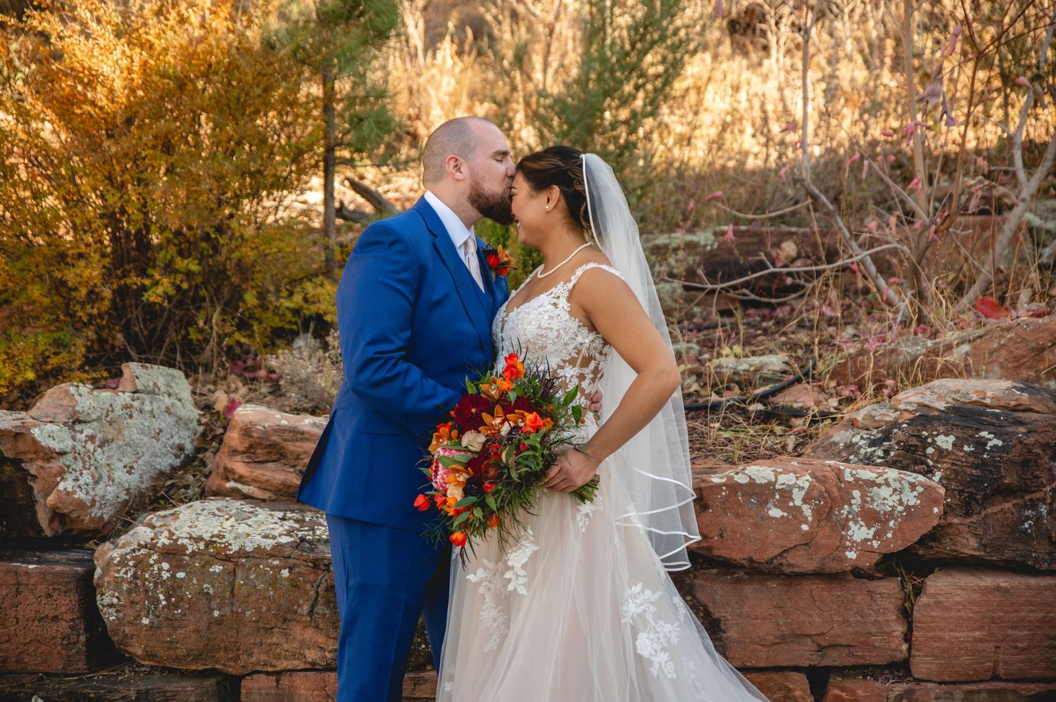 A romantic wedding photo of a couple in a blue suit and white dress against a rustic stone backdrop.