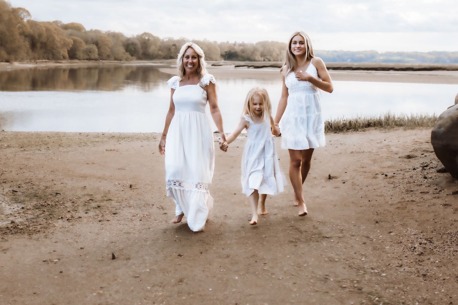 Two women and young girl in white dresses walking by lakeside together.