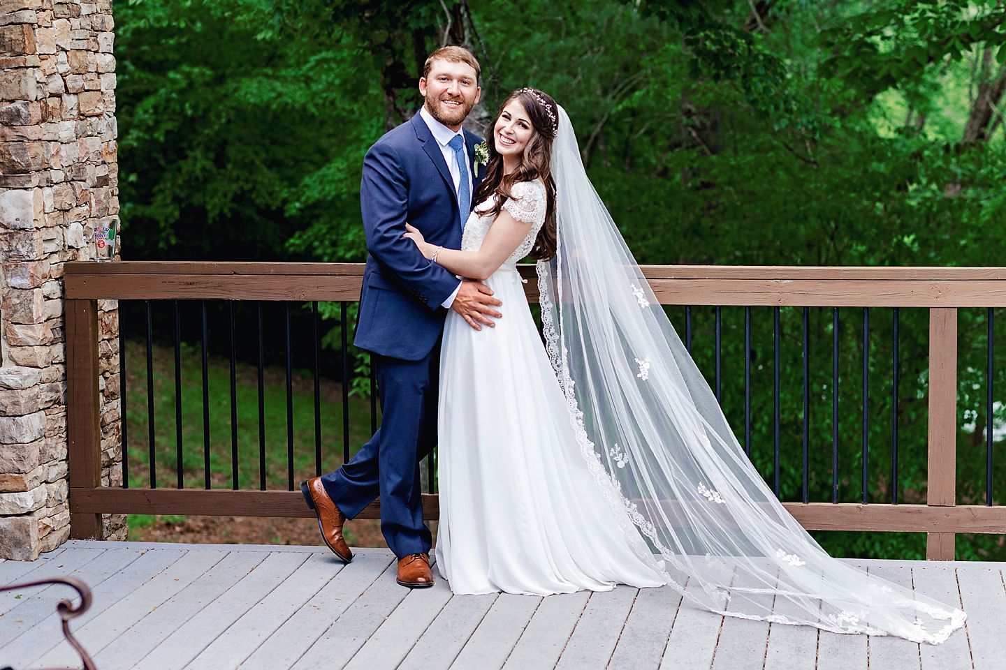 Newlyweds pose on wooden deck with bride's flowing veil and forest backdrop.