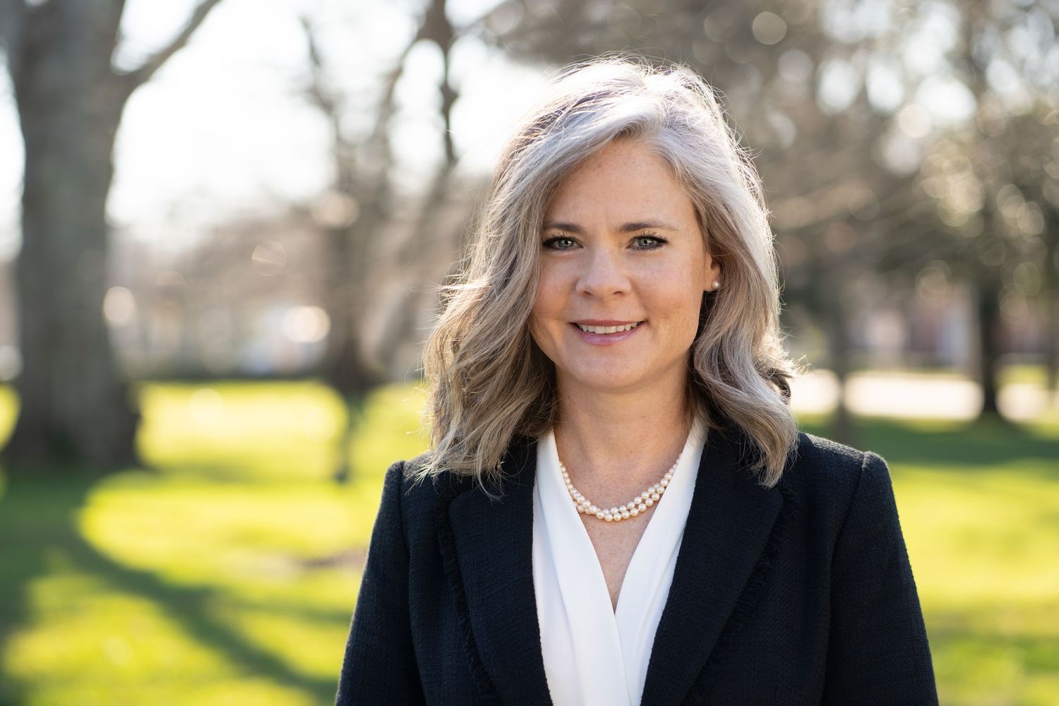 Professional headshot of woman in black blazer and pearl necklace outdoors.