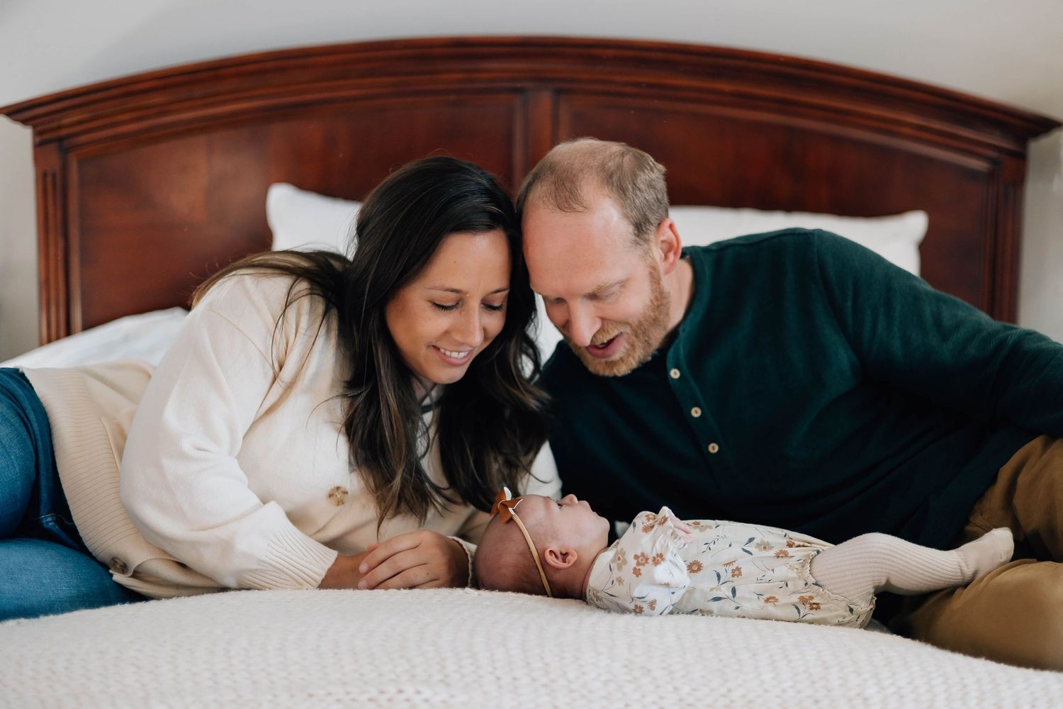 Smiling parents lying on a bed, gazing lovingly at their newborn baby between them.