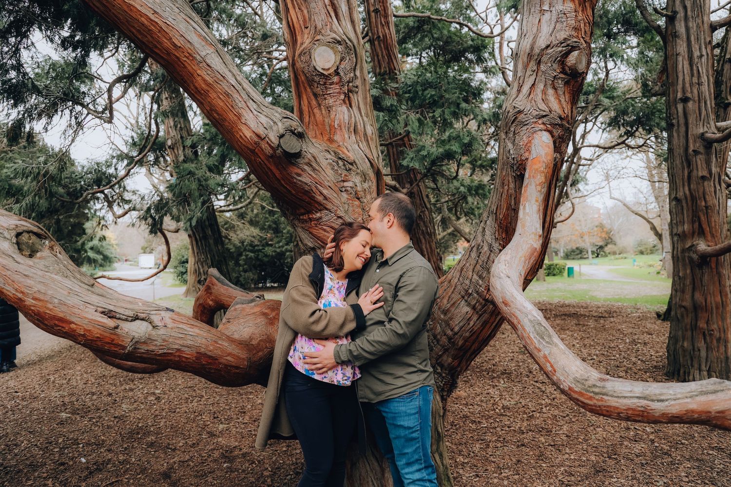 Romantic couple embracing near twisted tree trunks in a park setting.
