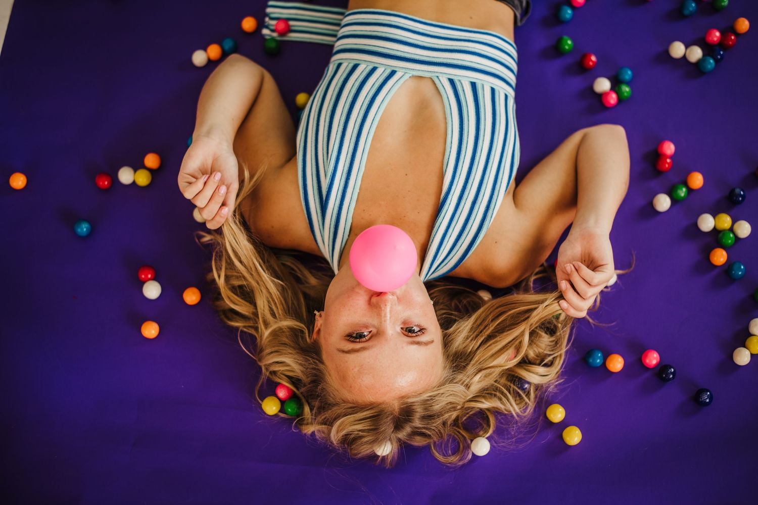 Overhead creative portrait with pink bubblegum and scattered colorful balls on purple surface.