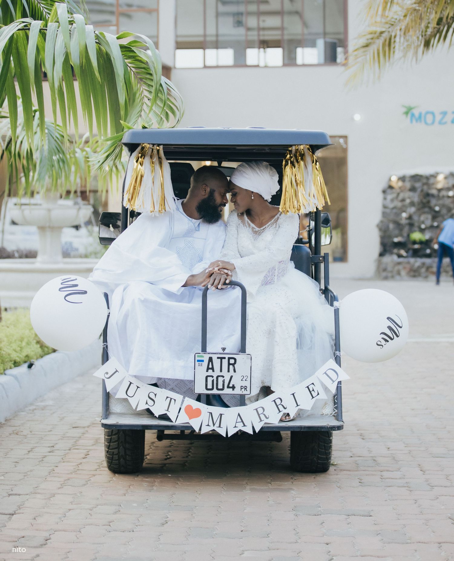 A newlywed couple in white attire sitting in a decorated golf cart with a Just Married sign.