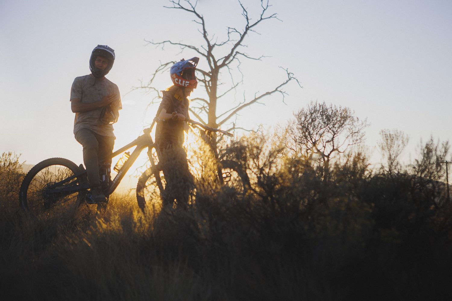 Two cyclists pause with their bikes silhouetted against a dramatic sunset with bare trees in the background.
