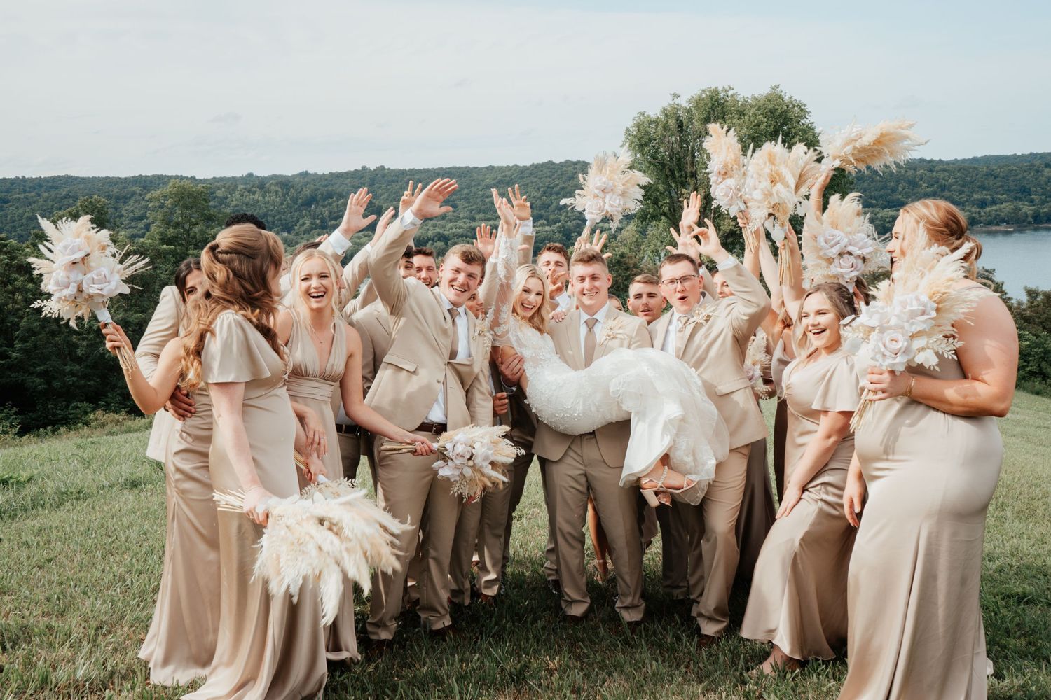 A wedding party celebrates outdoors holding pampas grass bouquets against a scenic backdrop.