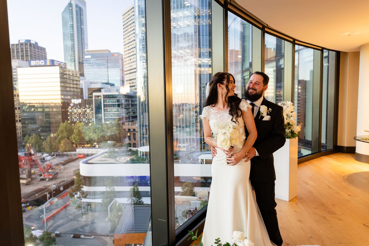 Newlywed couple poses against cityscape view through modern high-rise windows.