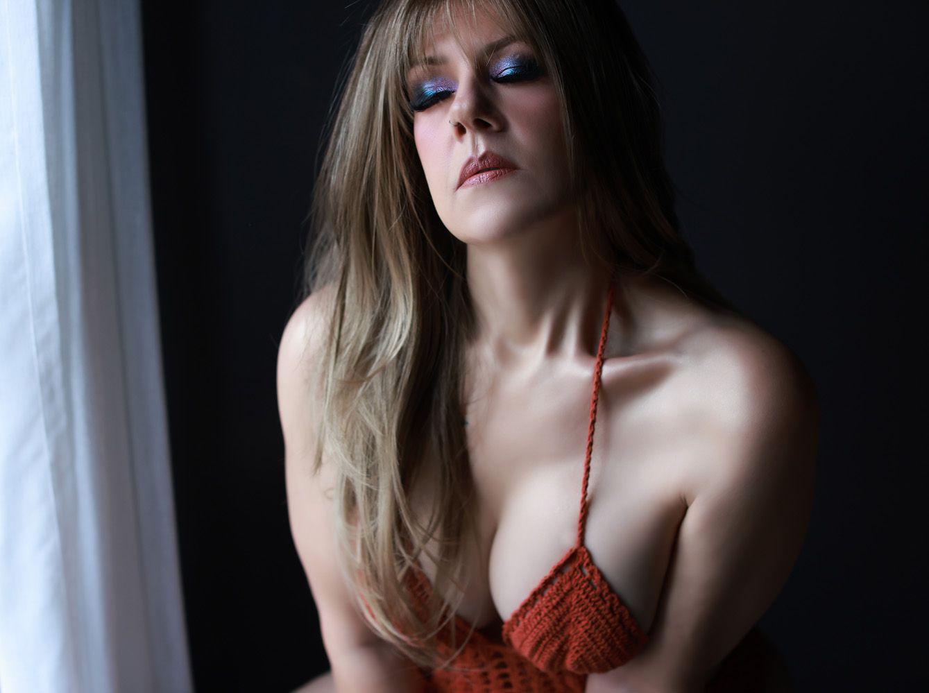 Woman with long blonde hair in red lingerie poses by window in moody lighting.
