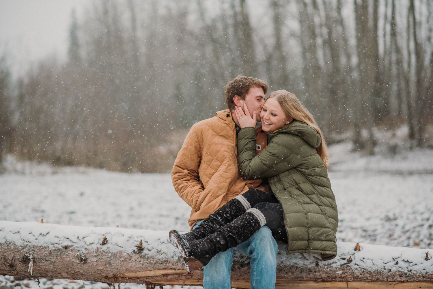 Couple Sitting on log, in snowy weather. Man is kissing girl on the face, girl has legs on mans lap
