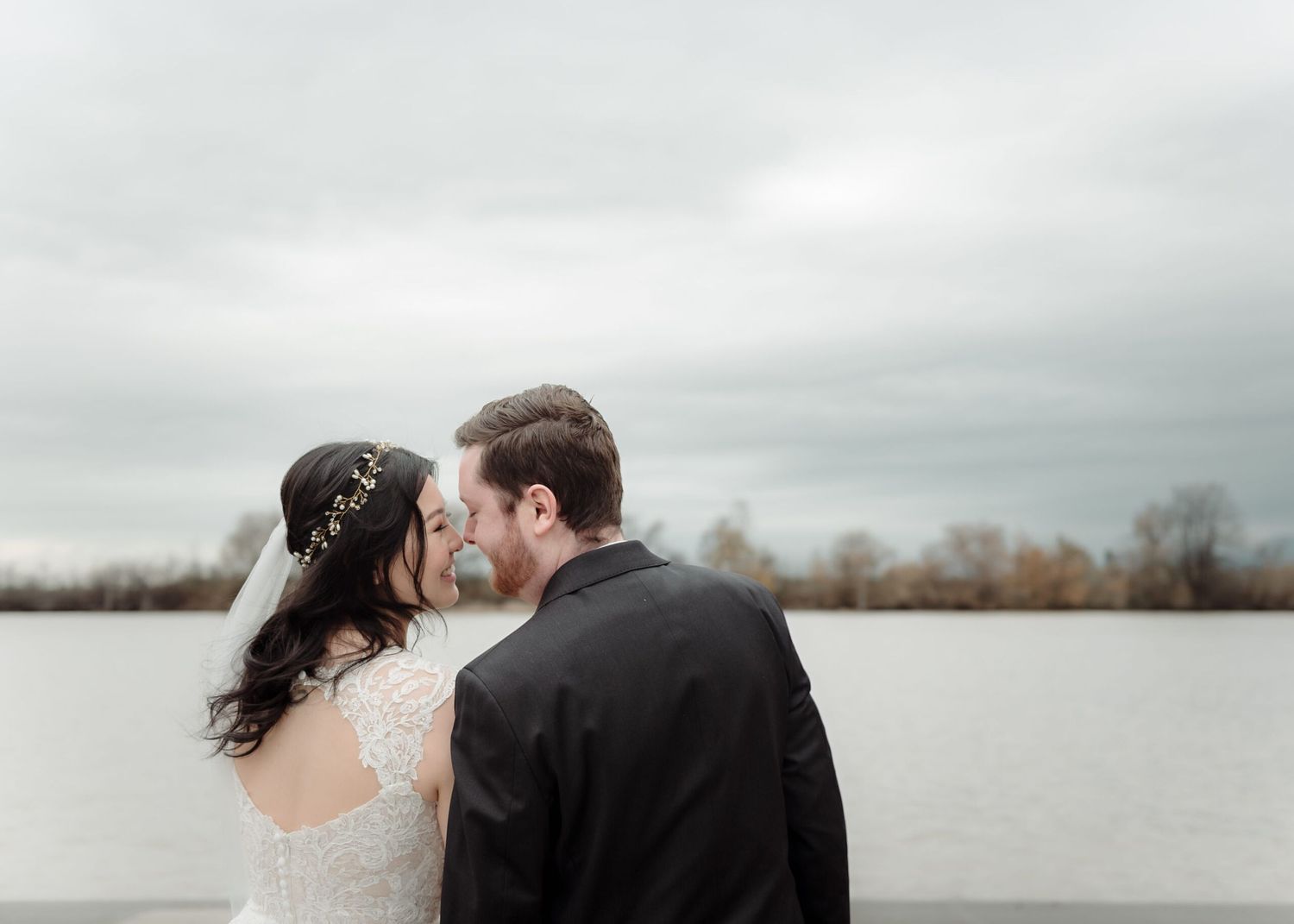 A romantic wedding couple embracing near a lake on an overcast day.