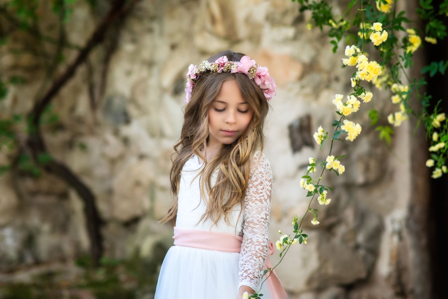 A white lace dress with long sleeves and floral crown captured against a stone wall with yellow flowers.