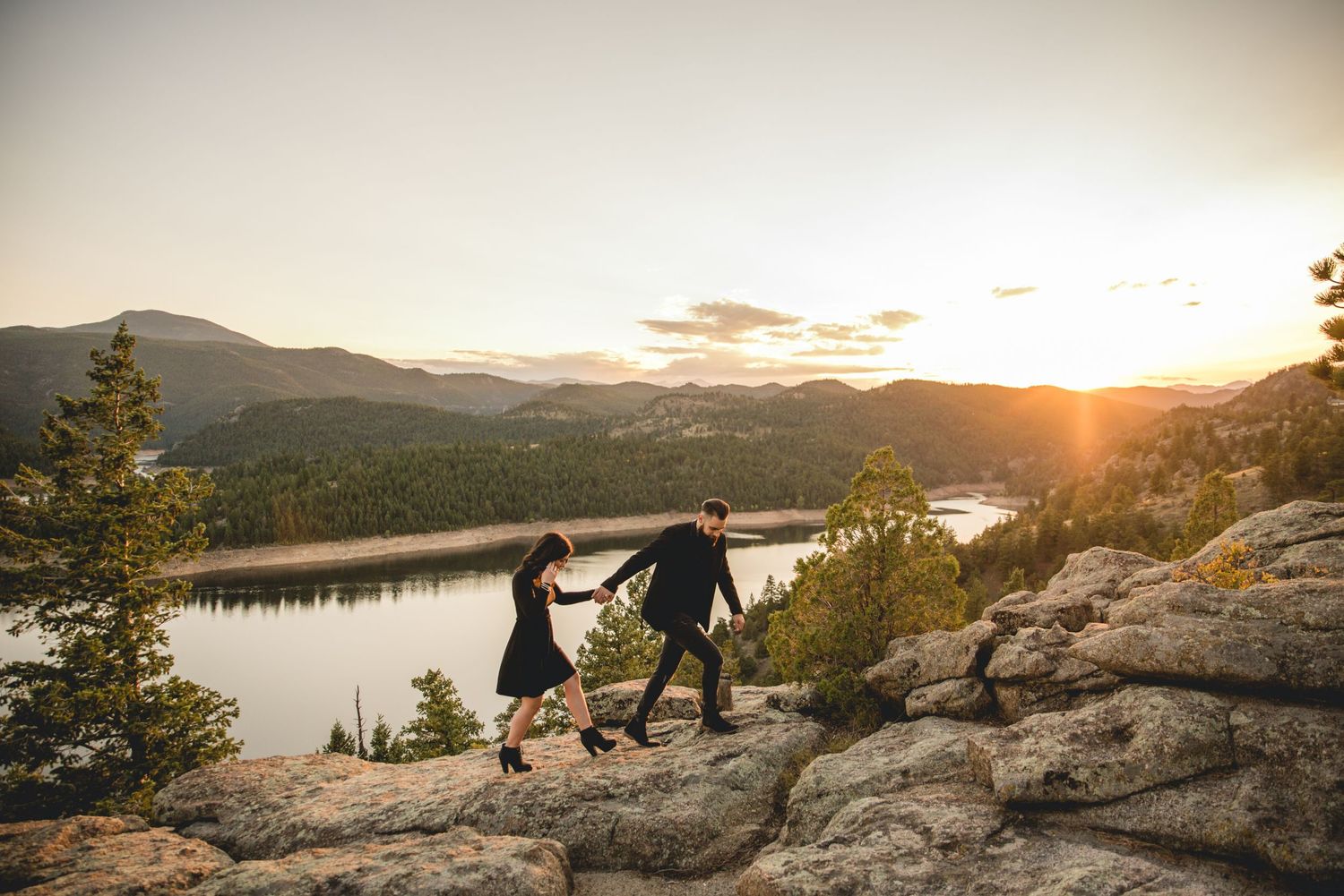 A couple holds hands while walking on rocky terrain at sunset near a mountain lake.