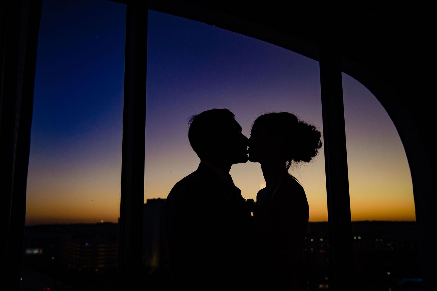 Silhouettes of a couple against a vibrant sunset sky through an arched window frame.