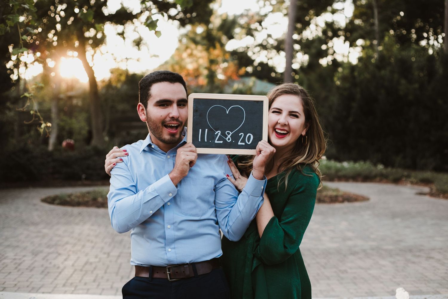 A couple holding a chalkboard sign with their wedding date outdoors at sunset.