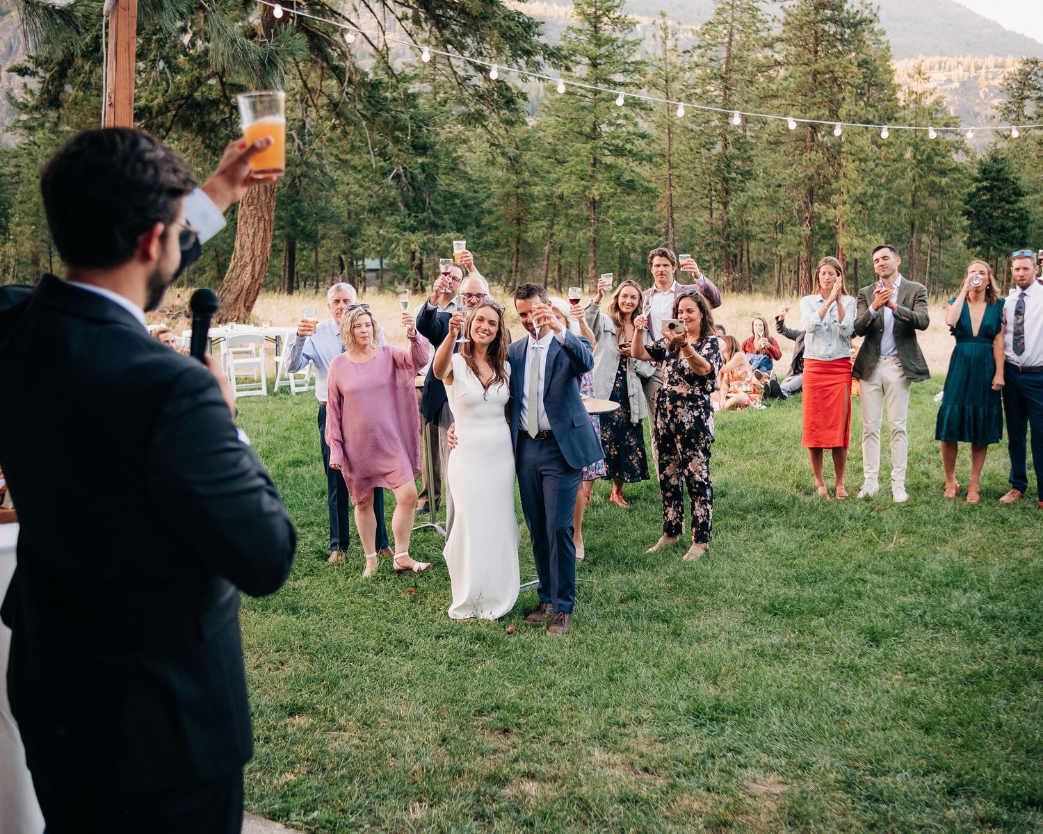 A group of wedding guests gather on a grassy lawn surrounded by pine trees during an outdoor celebration.