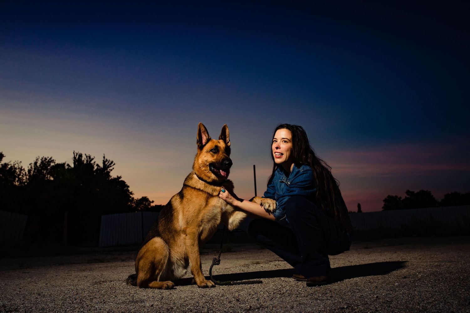 A German Shepherd dog poses with owner against a dramatic twilight sky.