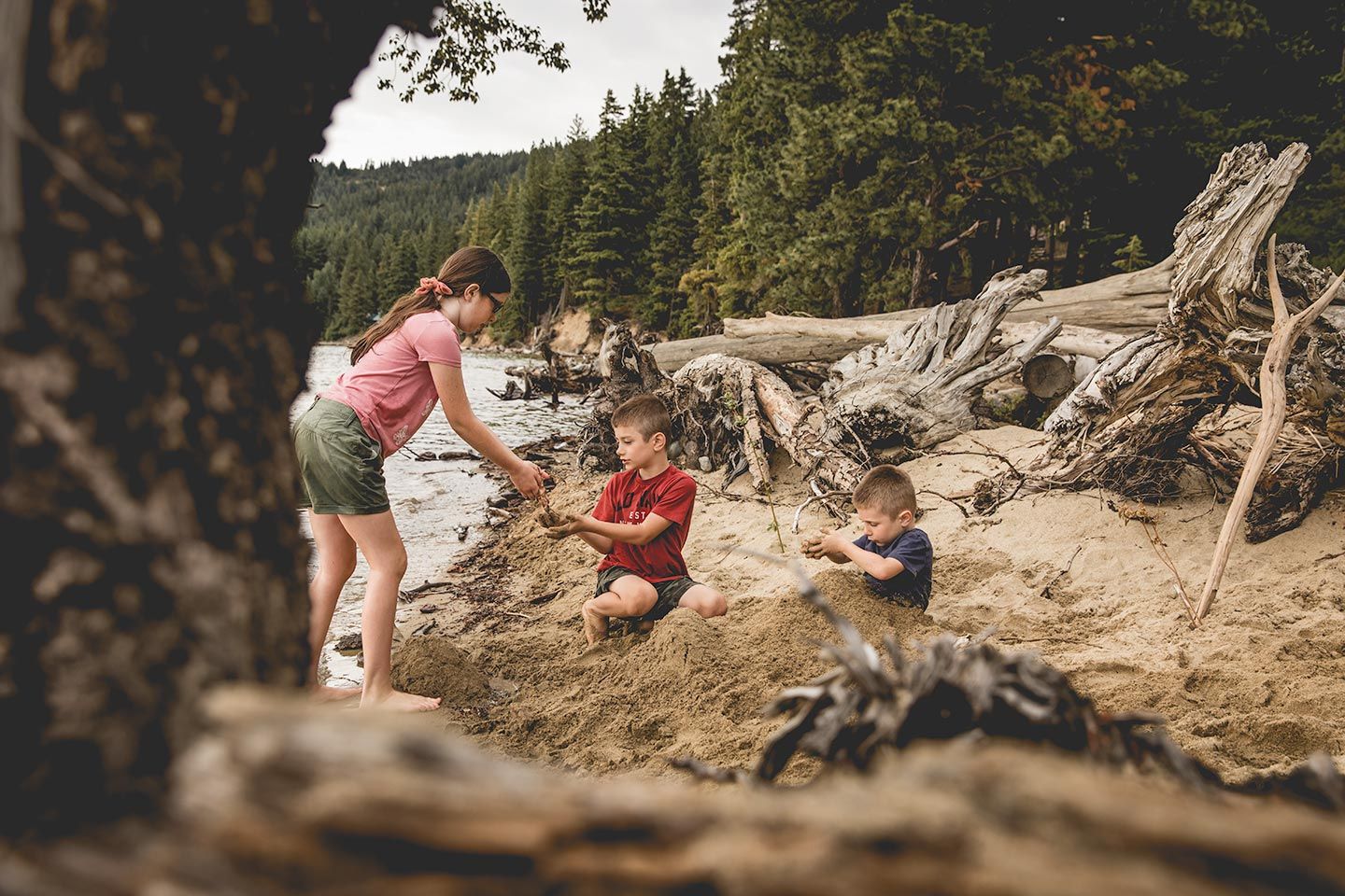 three kids playing in the sand at Cle Elum Lake. Photographer, Brindy Ruby, creates a unique perspective with the logs