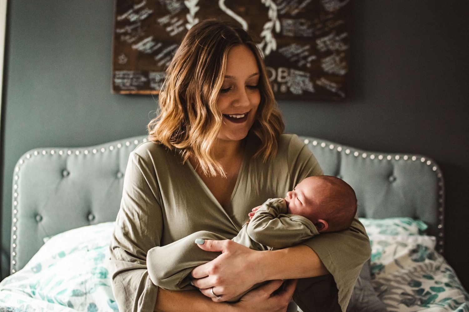 A tender moment showing a parent cradling a newborn baby on a gray tufted couch in natural lighting.