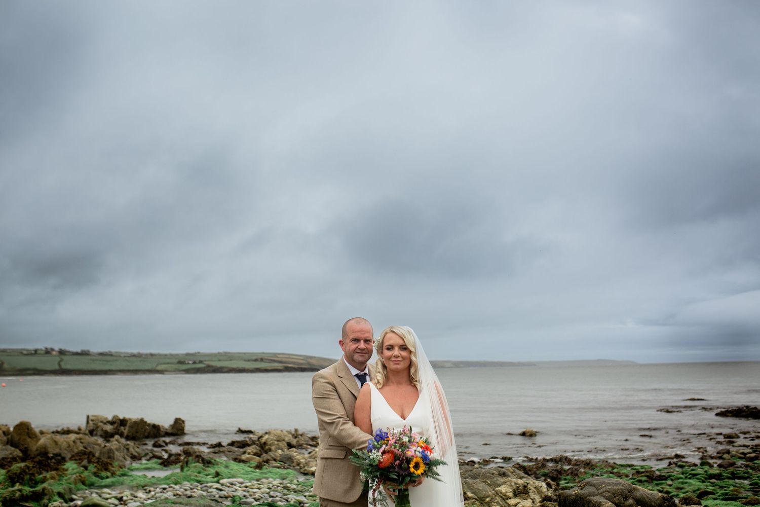 Newlyweds standing together on a cloudy beach with bouquet and formal attire.