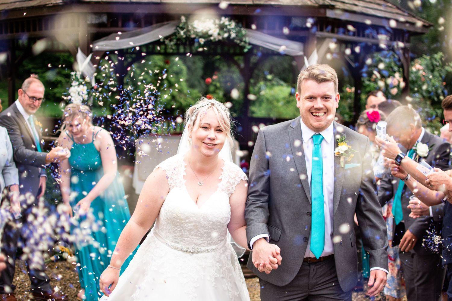 Wedding couple walks through shower of confetti as guests celebrate at outdoor ceremony.