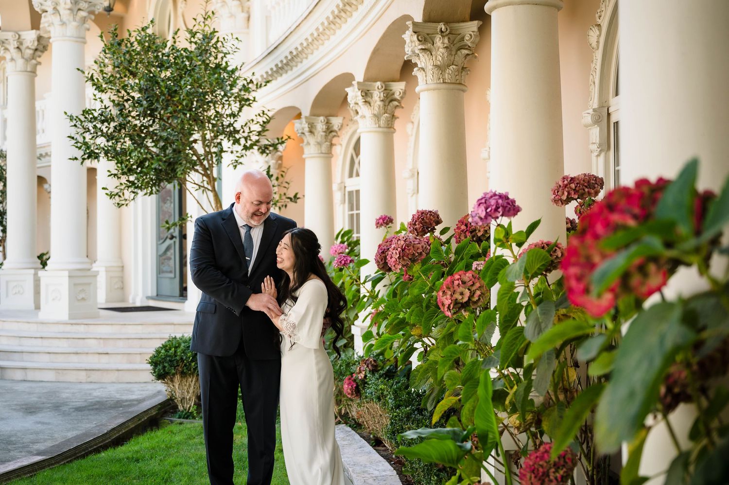 A couple posing by elegant classical columns with vibrant pink flowers in the foreground.