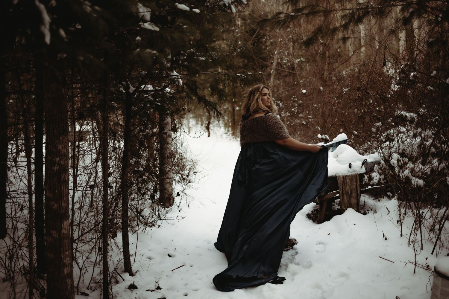 A person in a flowing dark dress poses dramatically in a snowy forest setting.