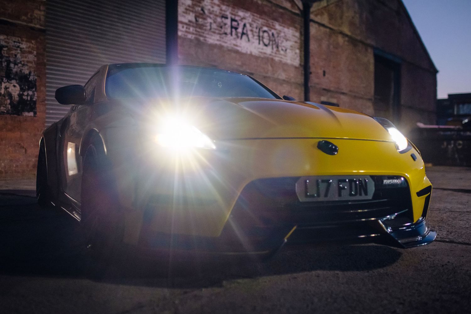 A yellow sports car with bright headlights photographed at sunrise against an industrial backdrop.