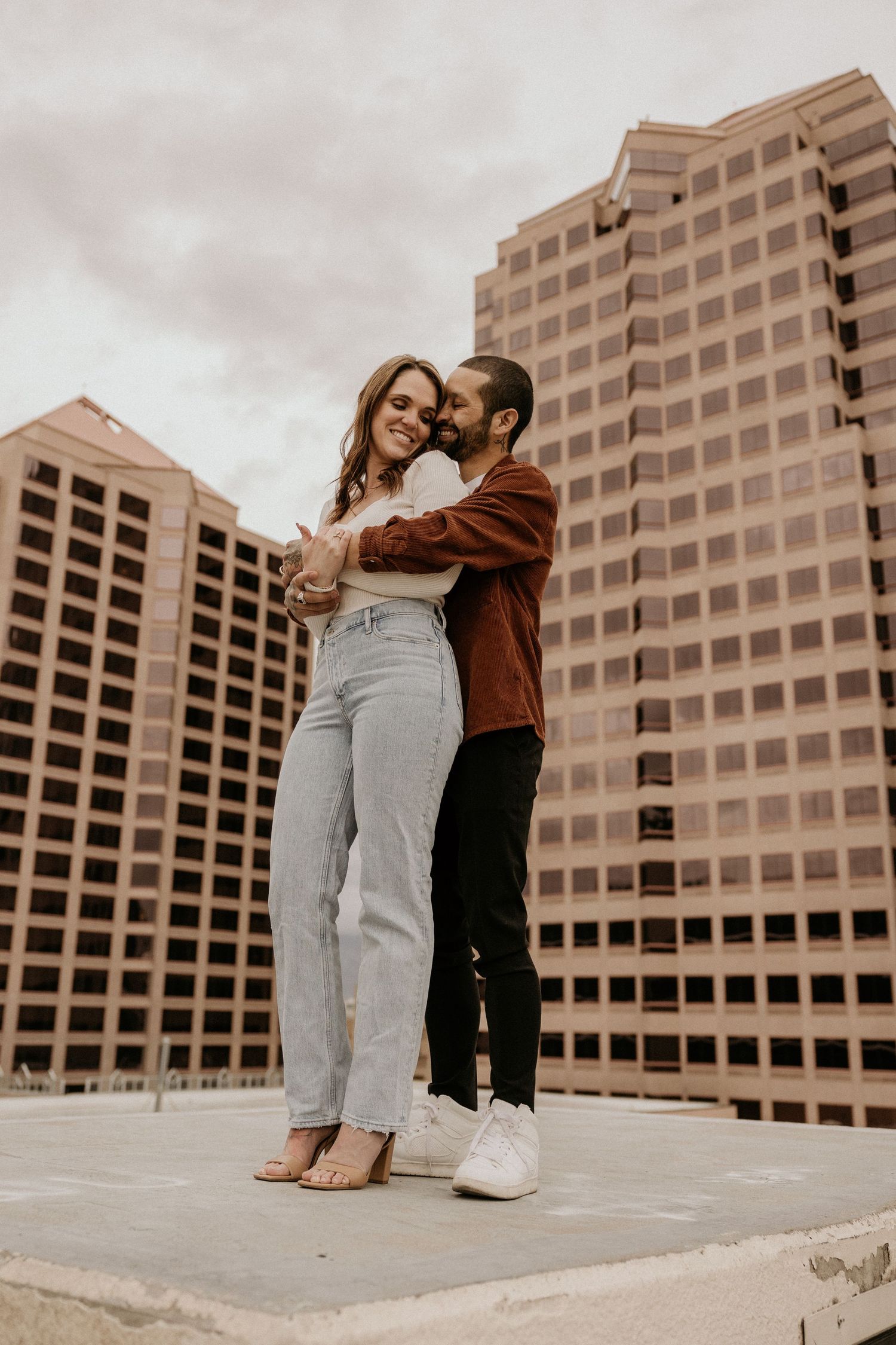 A couple embraces and smiles in front of tall city buildings on a rooftop.