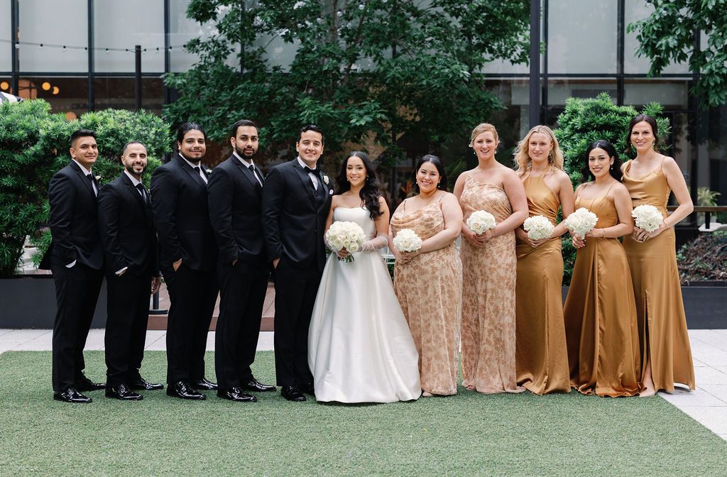 Wedding party in formal attire poses together on artificial turf with bridesmaids in gold sequin dresses.