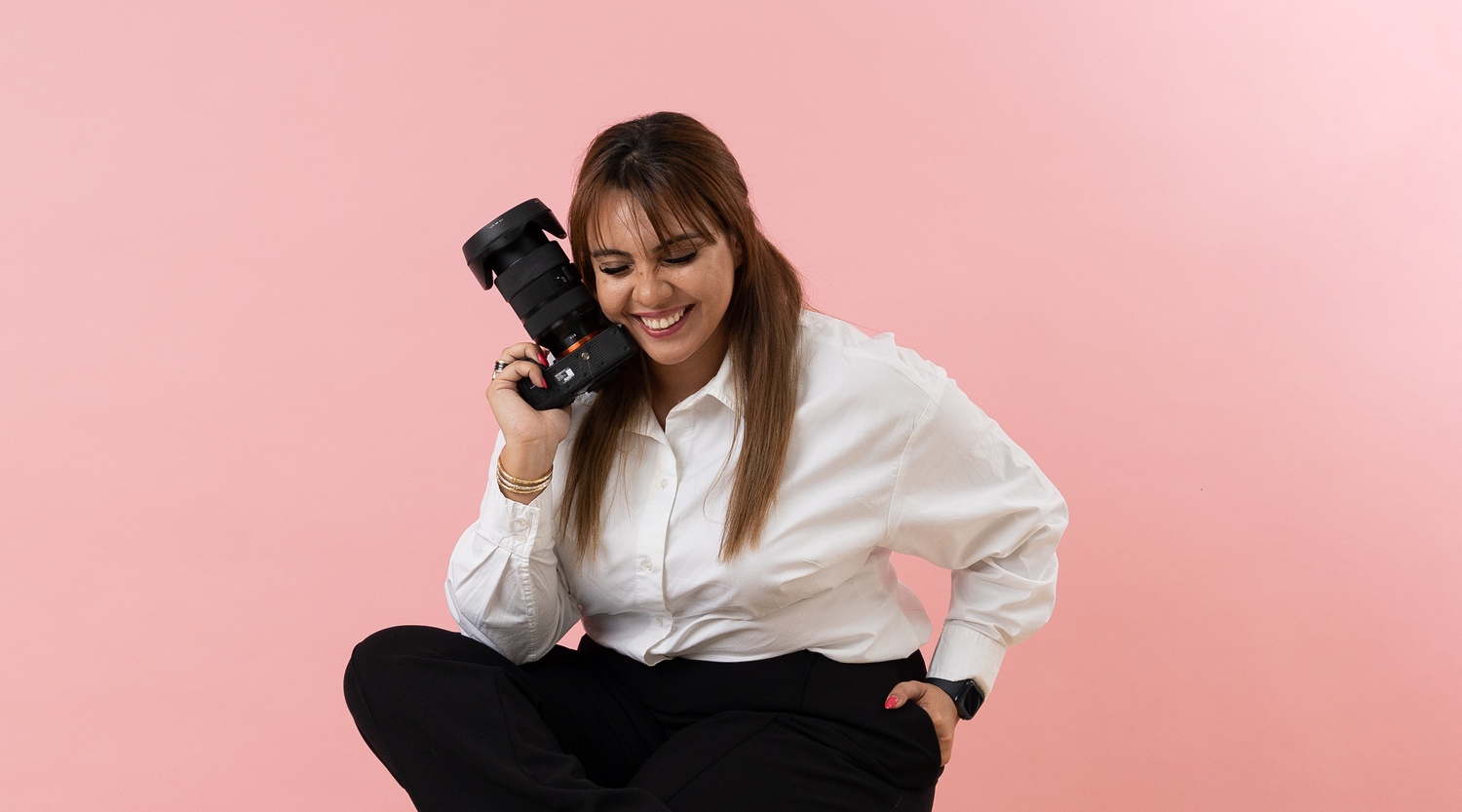 Professional photographer in white button-down shirt and black pants crouches with camera against pink backdrop.