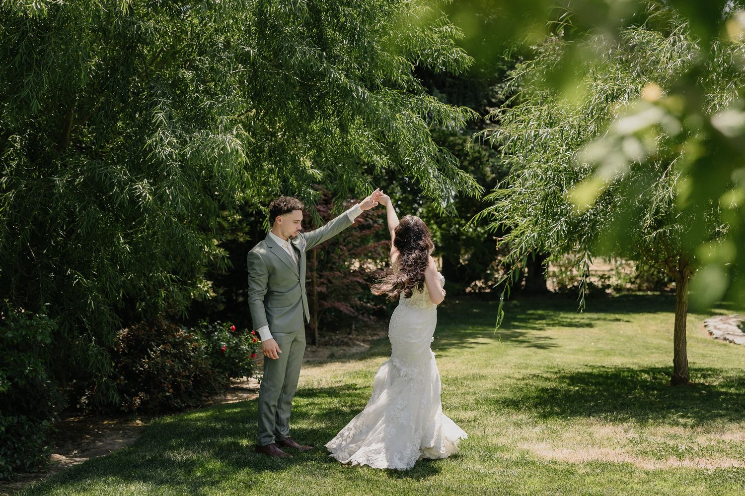 A couple dances together on a sunny summer day in a lush garden setting with overhanging trees.