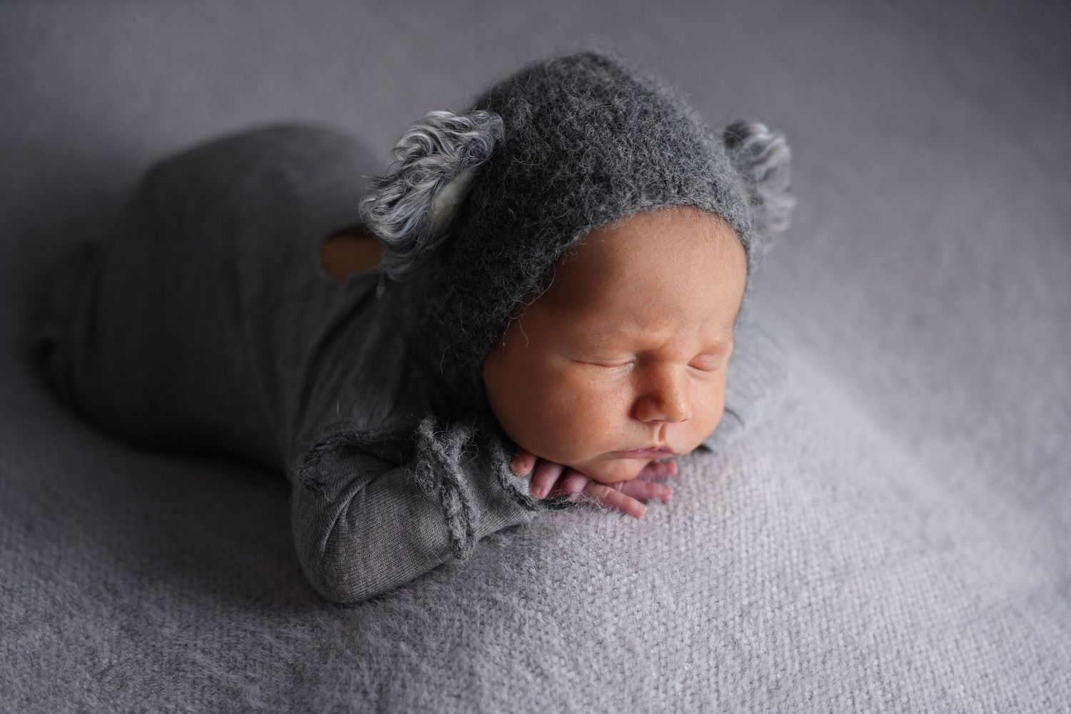 A peaceful sleeping newborn wearing a cozy gray bear outfit lays on light gray textured fabric.