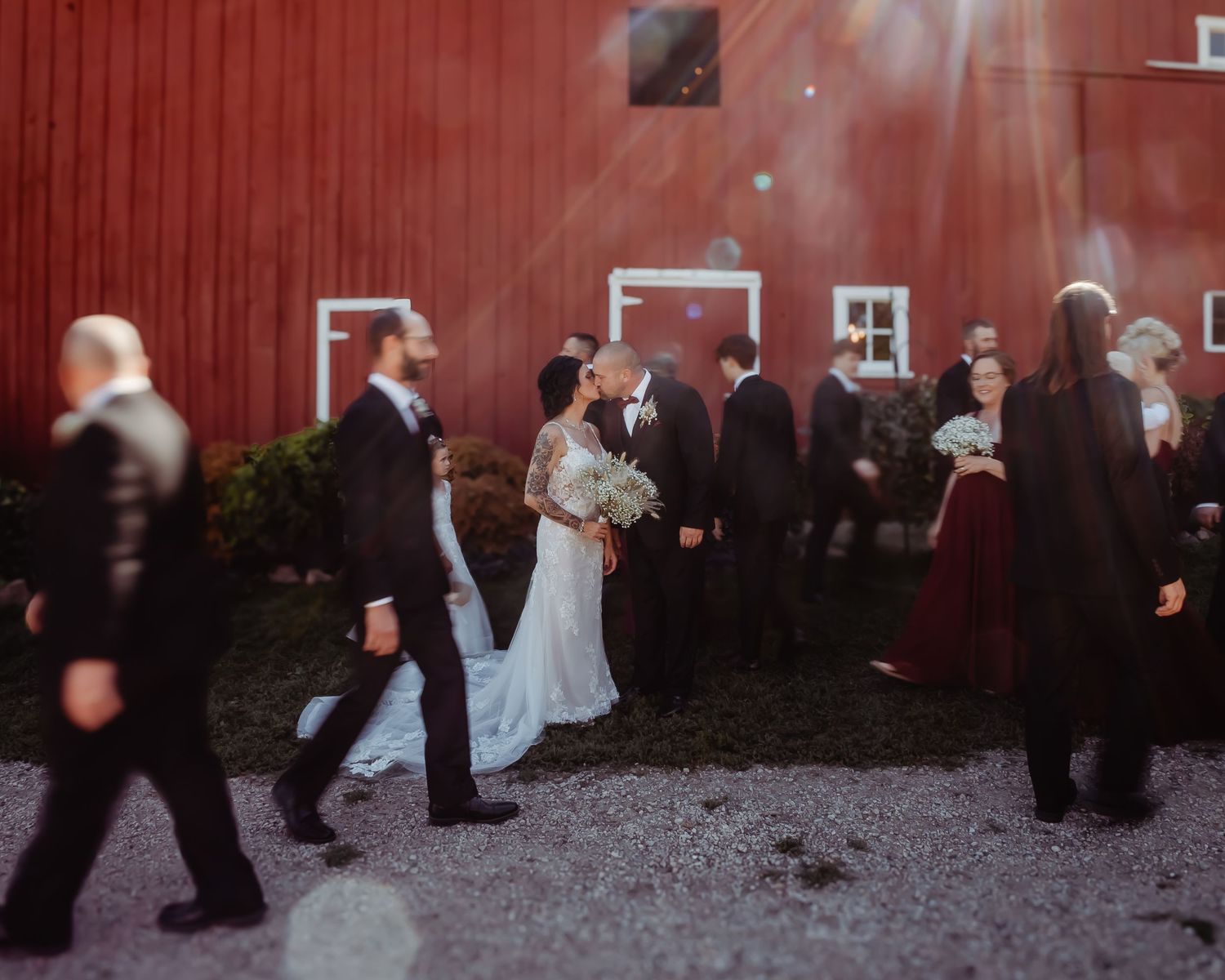 People in formal attire gather outside a rustic red barn during an outdoor wedding celebration with misty light streaming down.