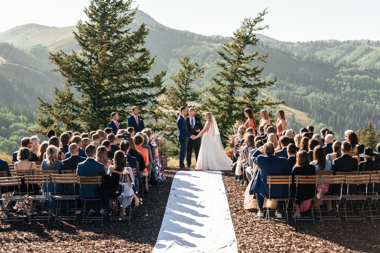 A Park City mountain wedding ceremony takes place outdoors with guests seated on wooden benches overlooking a scenic mountain landscape.