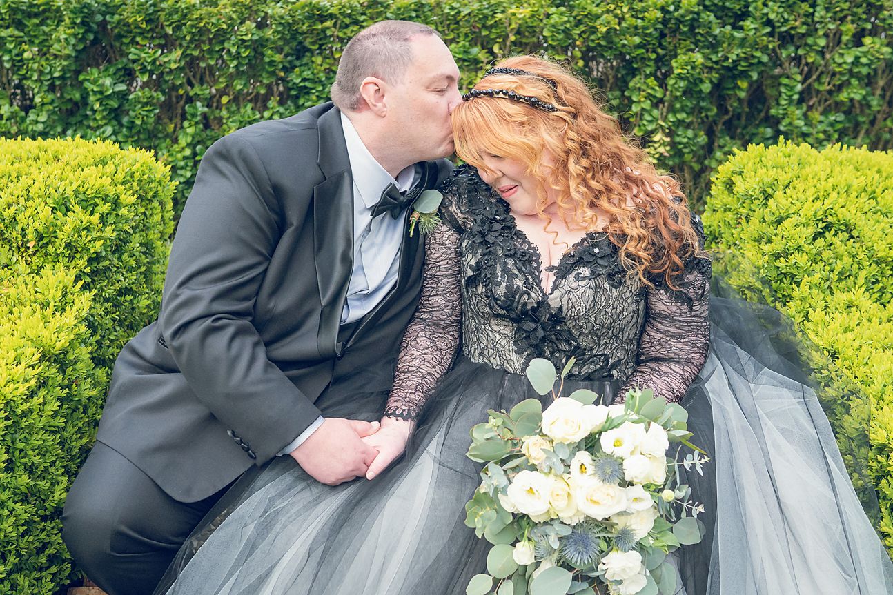 A newlywed couple dressed in grey and black shares romantic kisses in a formal hedge garden on their Hepburn Springs wedding day.