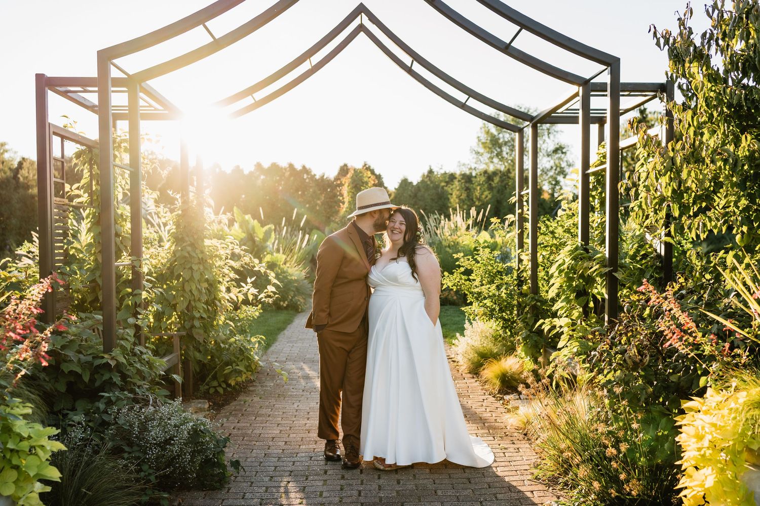 A groom whispers in his bride's ear in front of a garden structure at sunset.