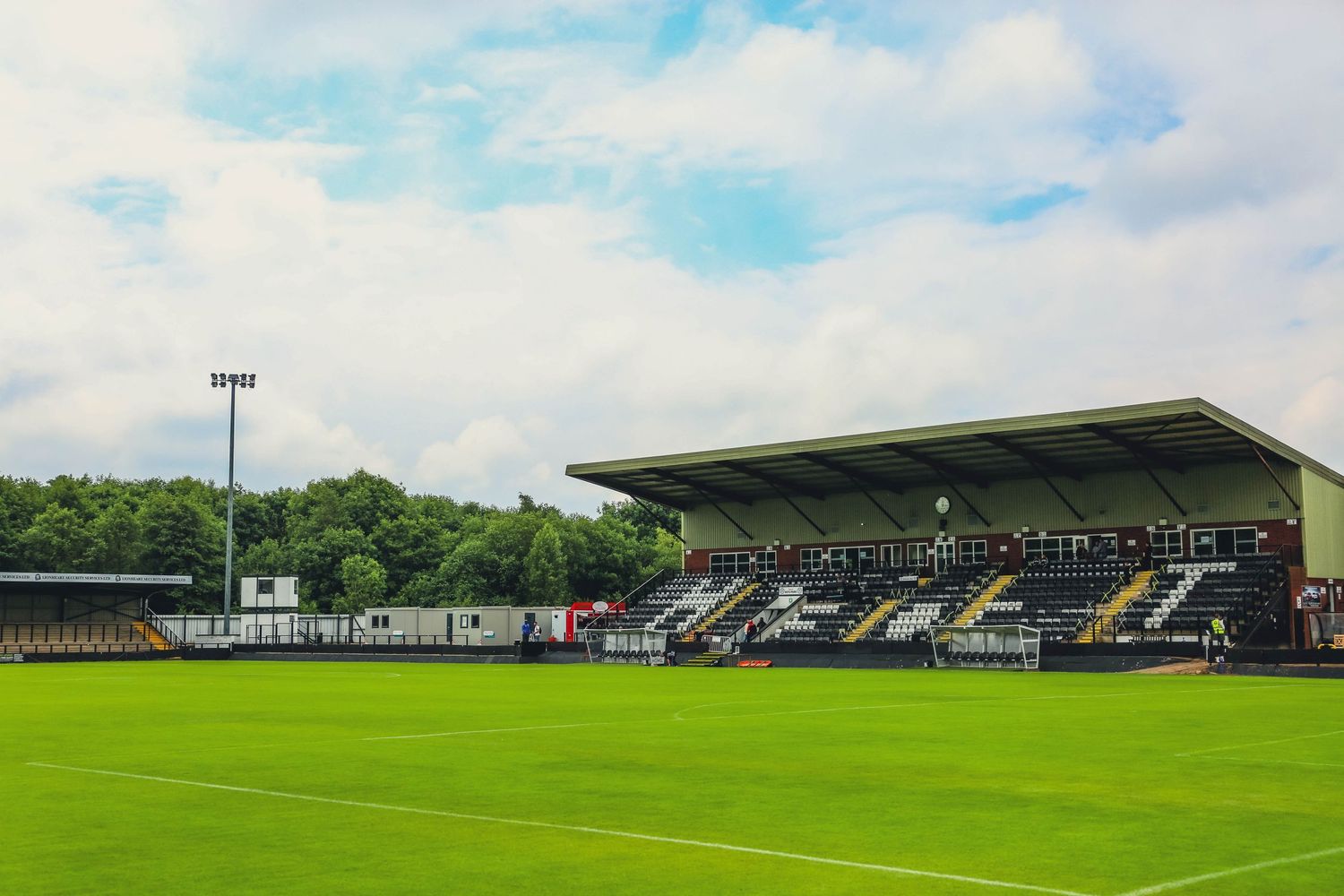 Wide view of an empty football stadium with green pitch and covered grandstand under blue sky.