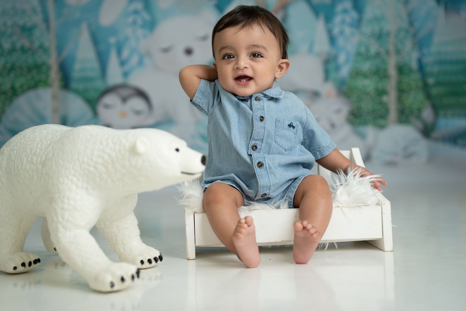 Little one sits on white wooden bench beside large decorative polar bear against dreamy blue backdrop.