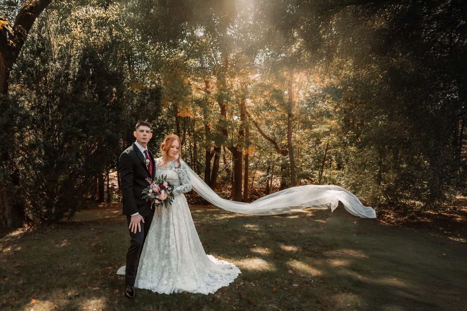 A couple in wedding attire poses in a sunlit forest as the bride's veil flows dramatically in the wind.