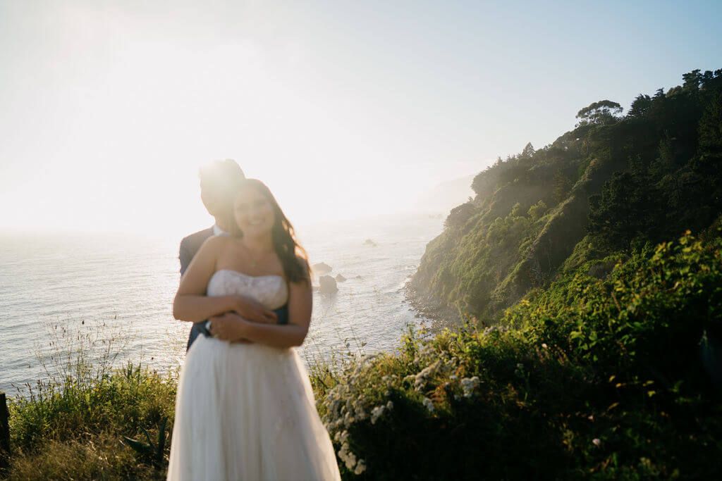 Wedding ceremony guests seated in rows at an outdoor cliffside venue overlooking the blue ocean under tall trees.