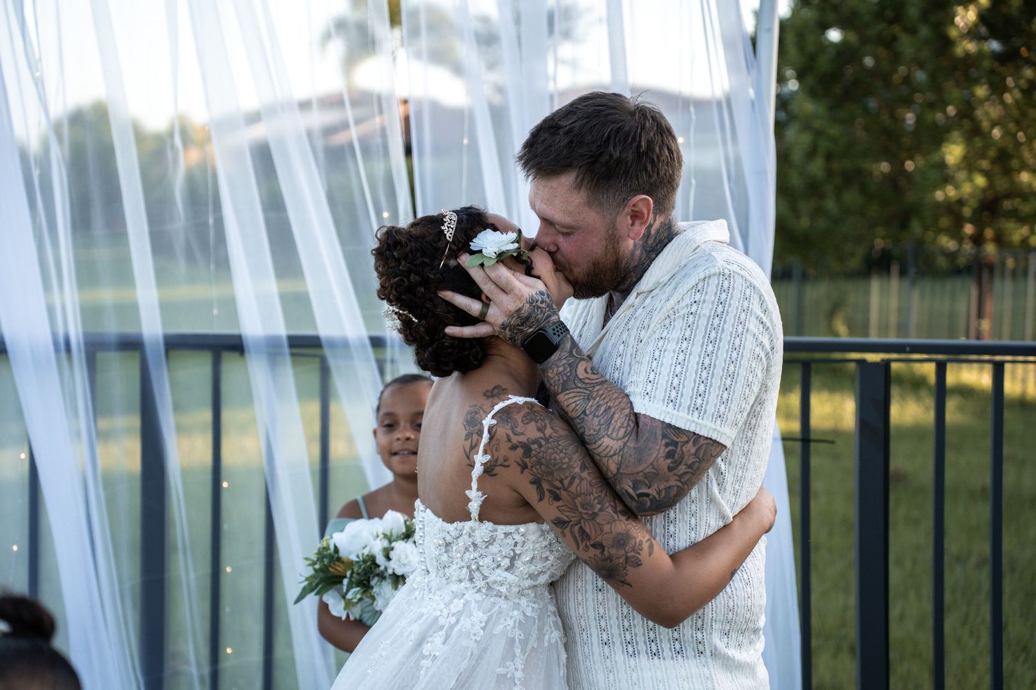 A serene bride in a flowing white dress poses against a rustic wooden fence in a sunlit outdoor garden setting.