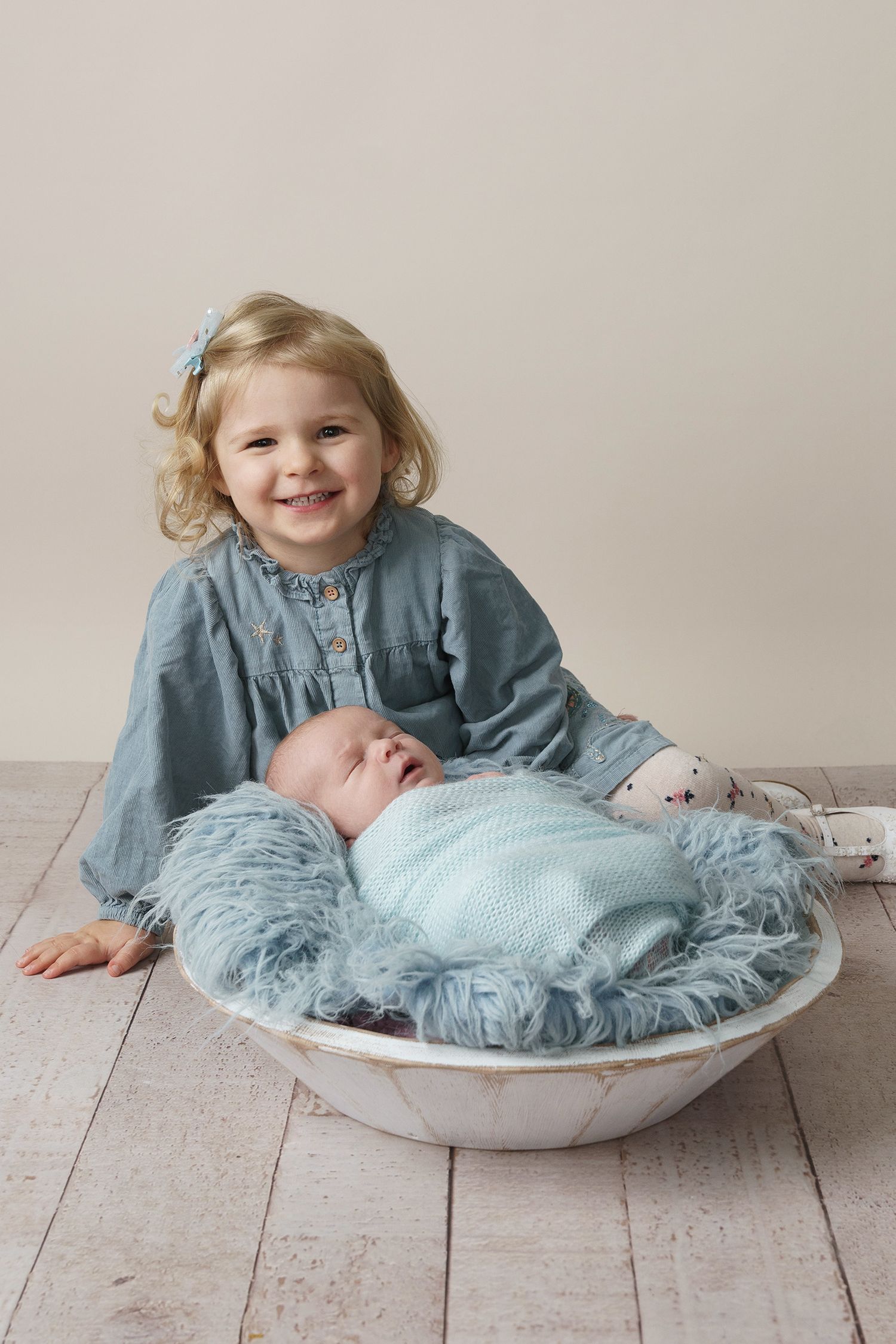 Two children in soft blue outfits sit together in a white bowl on a light wooden floor.