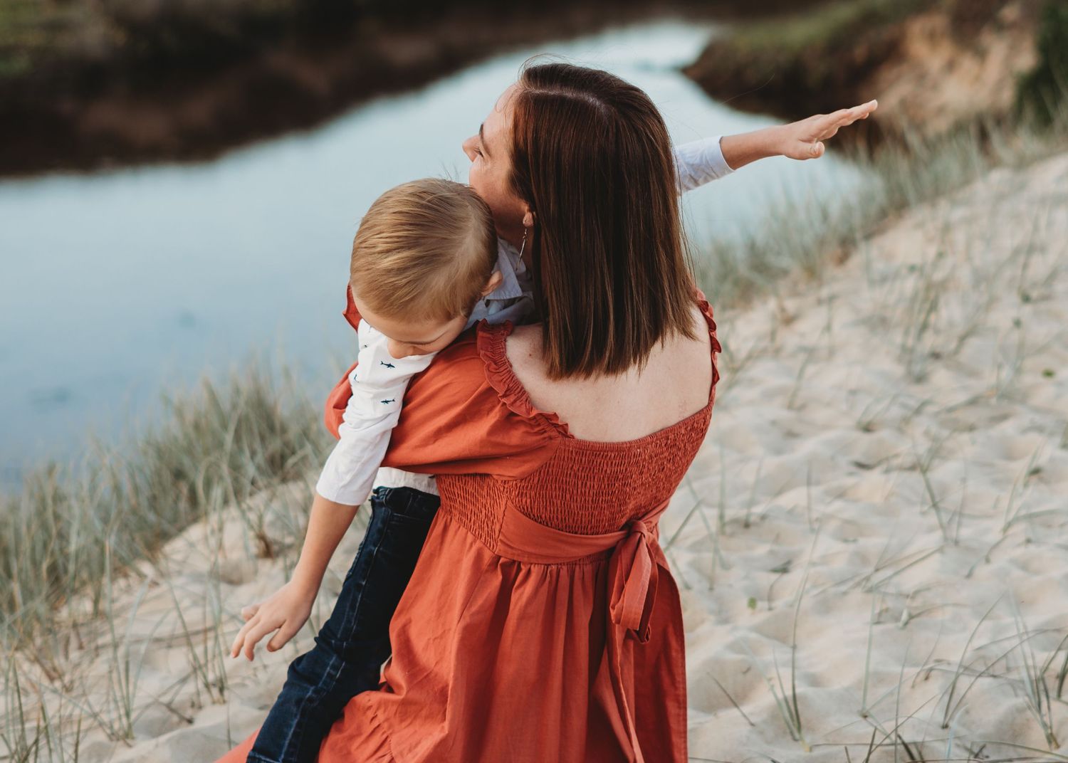 Mother in red dress holding toddler son while pointing at scenic sandy beach landscape.