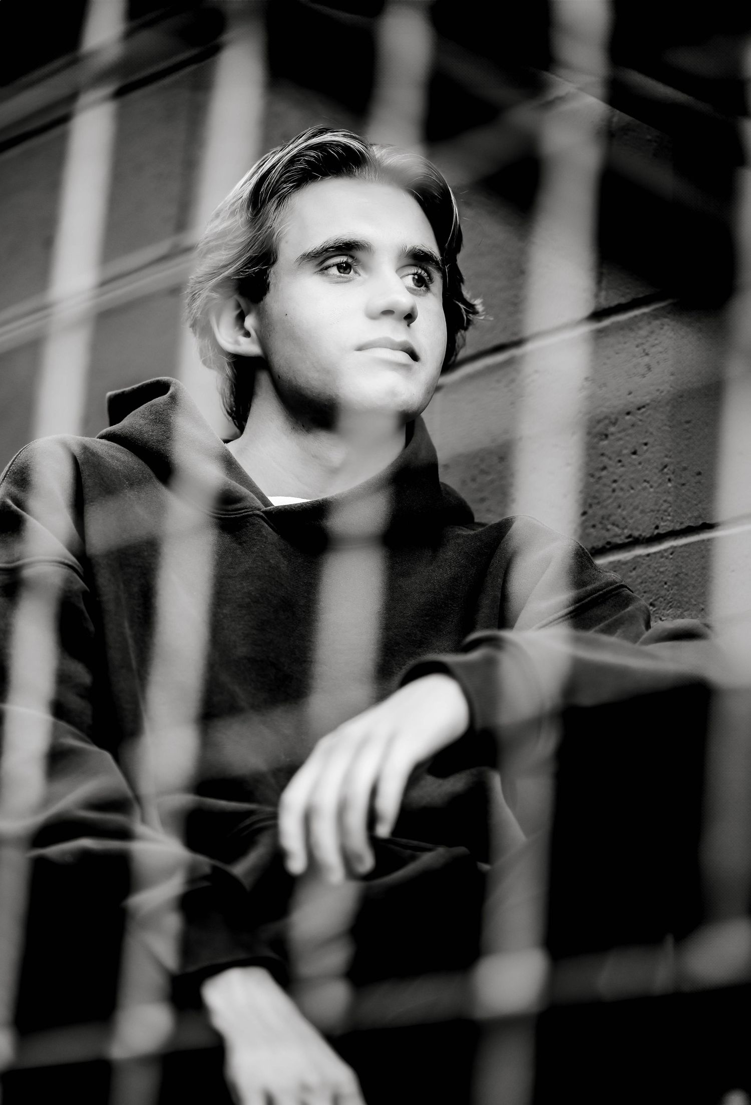 Black and white portrait of a young man looking up thoughtfully through blurred fence bars.
