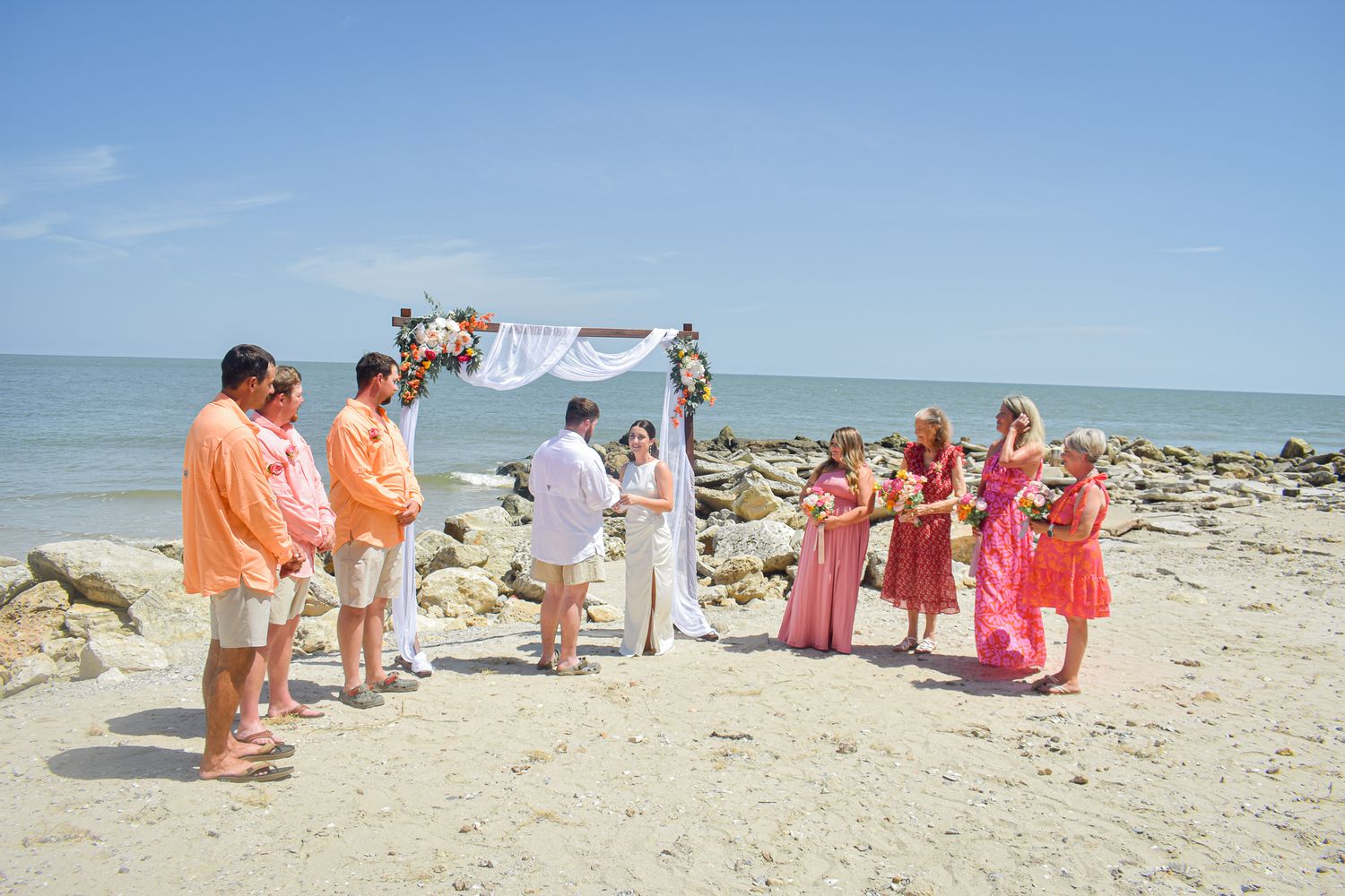 Wedding guests gather around a floral arch on a rocky beach during a seaside ceremony.