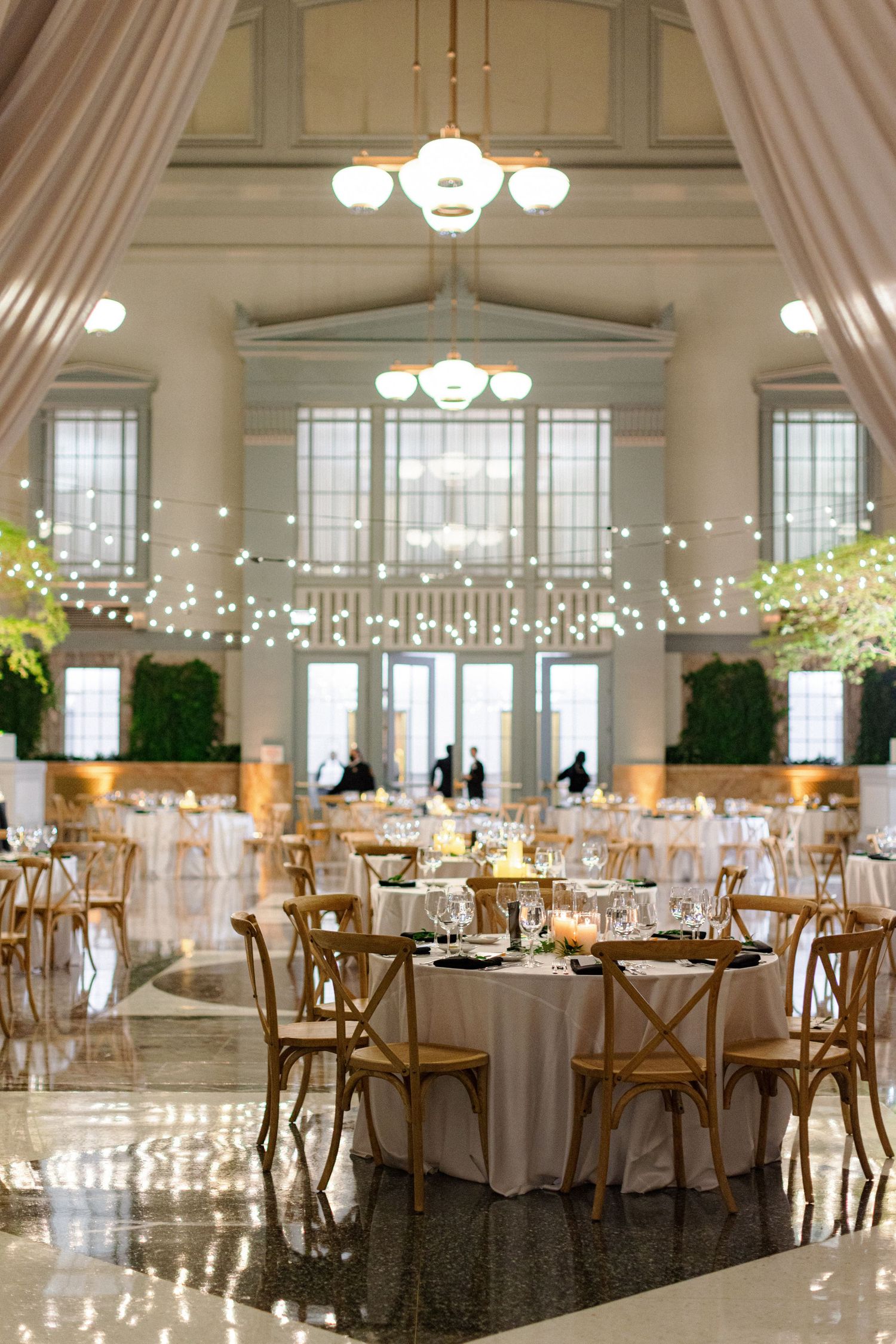 Elegant wedding reception venue with string lights, crystal chandeliers and rustic cross-back chairs around white-clothed tables.